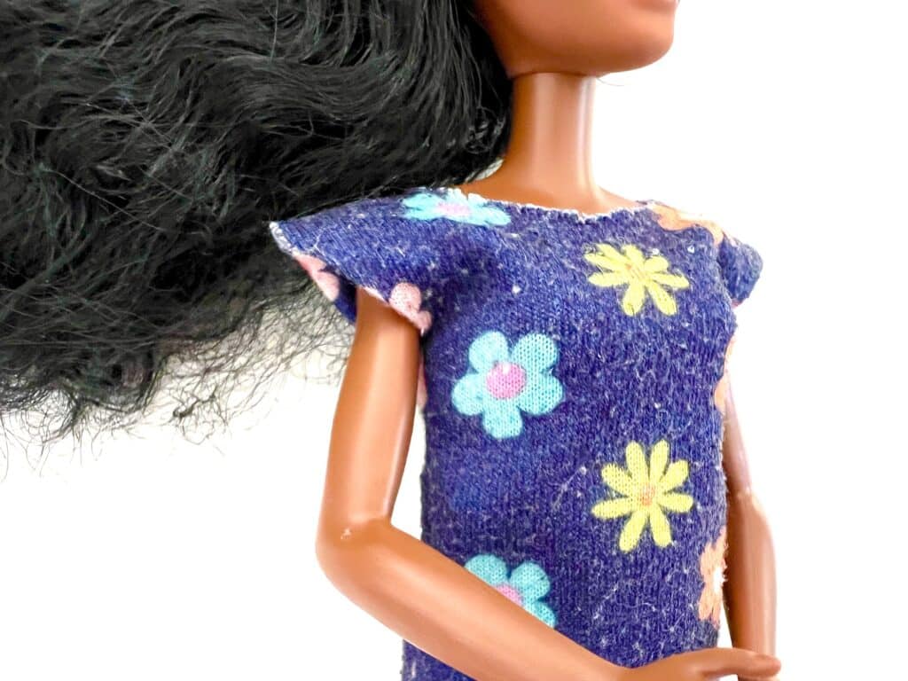 A doll with long dark hair, wearing a blue dress patterned with yellow and blue flowers, reminiscent of a classic Barbie shirt style.