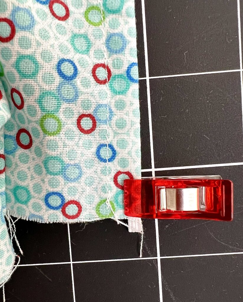 Close-up of a red sewing clip holding colorful, patterned fabric with hexagon designs. The fabric is placed on a black, grid-lined cutting mat.