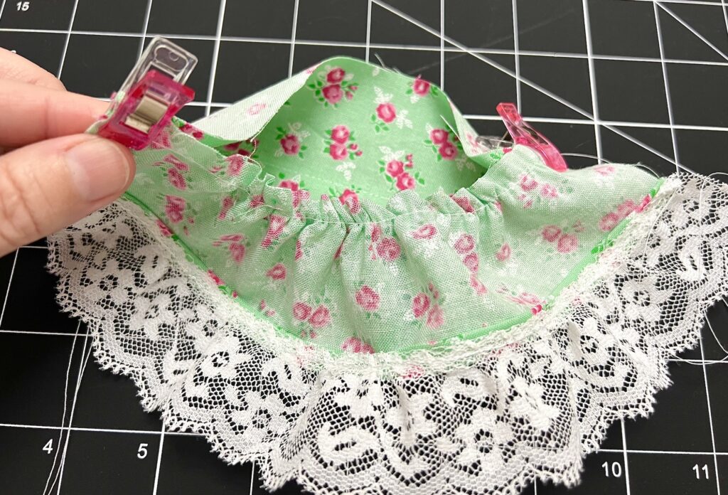 A hand holds a pink clip on green floral fabric with a gathered edge and white lace trim, reminiscent of a tiered skirt's elegant layers, all set against a black grid surface.