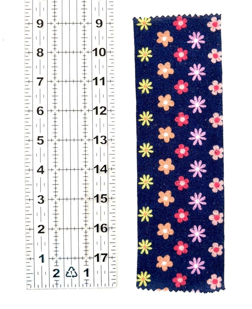 A measuring ruler lies beside a swatch of dark fabric adorned with a vibrant, multicolored floral pattern, reminiscent of a playful Barbie shirt.