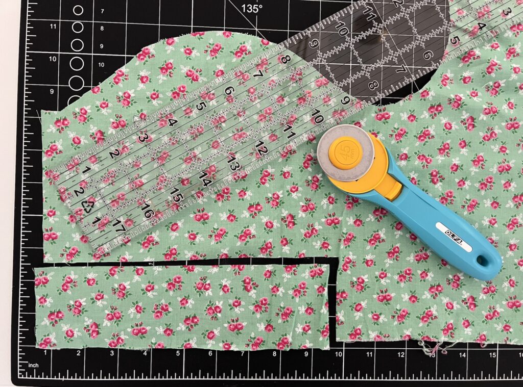 Green floral fabric lies on a cutting mat, ready to be crafted into a lovely tiered skirt. Nearby, a yellow and blue rotary cutter and transparent rulers await their turn to shape the vibrant material into something truly special.