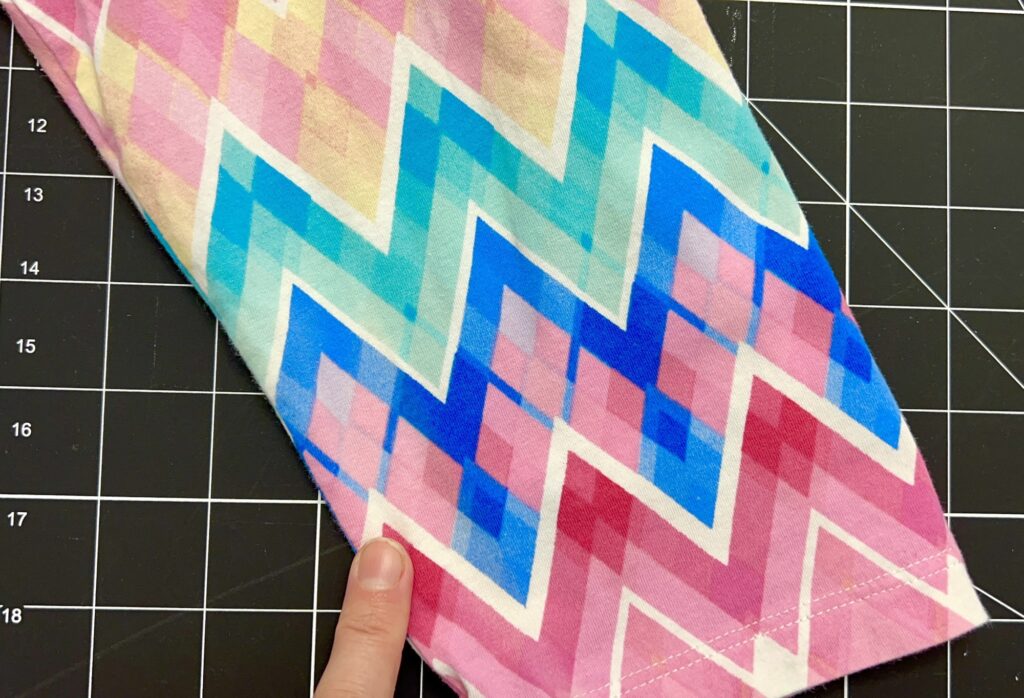 A finger points to a fabric with a zigzag pattern in pastel colors, primarily pink, blue, and beige, set against a black grid background.