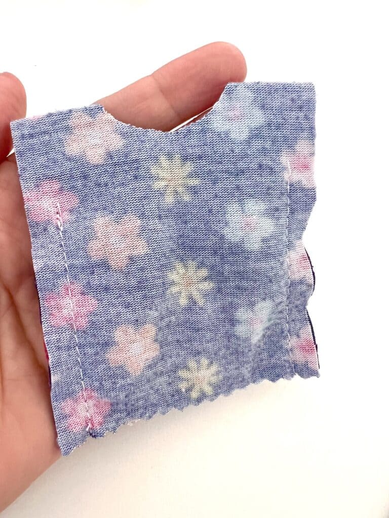 A hand delicately holds a small piece of blue fabric, adorned with pink and yellow floral patterns reminiscent of a Barbie shirt. The visible stitches offer a touch of crafted charm, all set against a pristine white background.