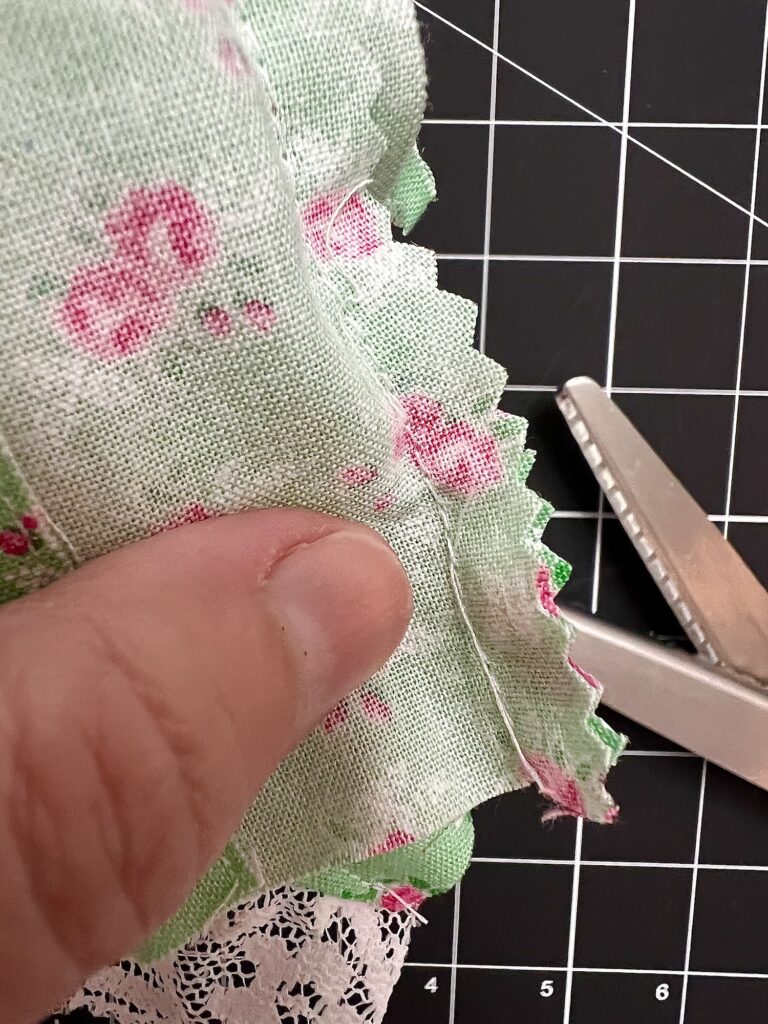 Close-up of a hand skillfully cutting green fabric adorned with pink flowers using pinking shears on a black cutting mat with measurement grid, laying the groundwork for a beautiful tiered skirt.