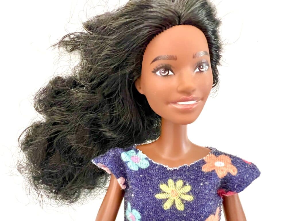 A fashion doll with long dark hair, wearing a colorful floral dress and a stylish Barbie shirt, smiling.