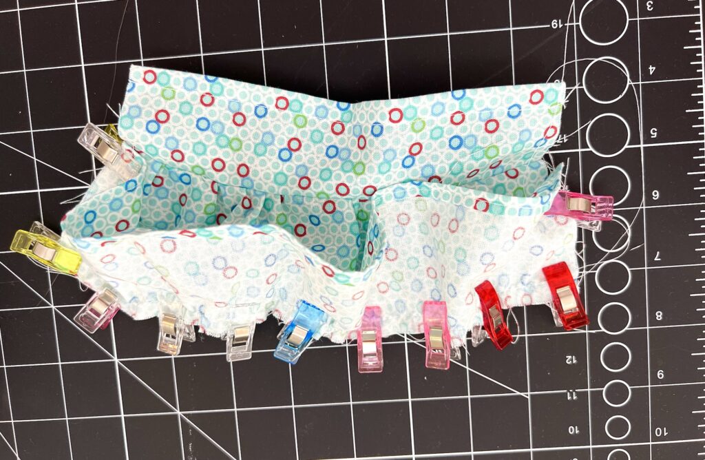 Fabric with multicolored circles is held with various clips on a black cutting mat with a grid.