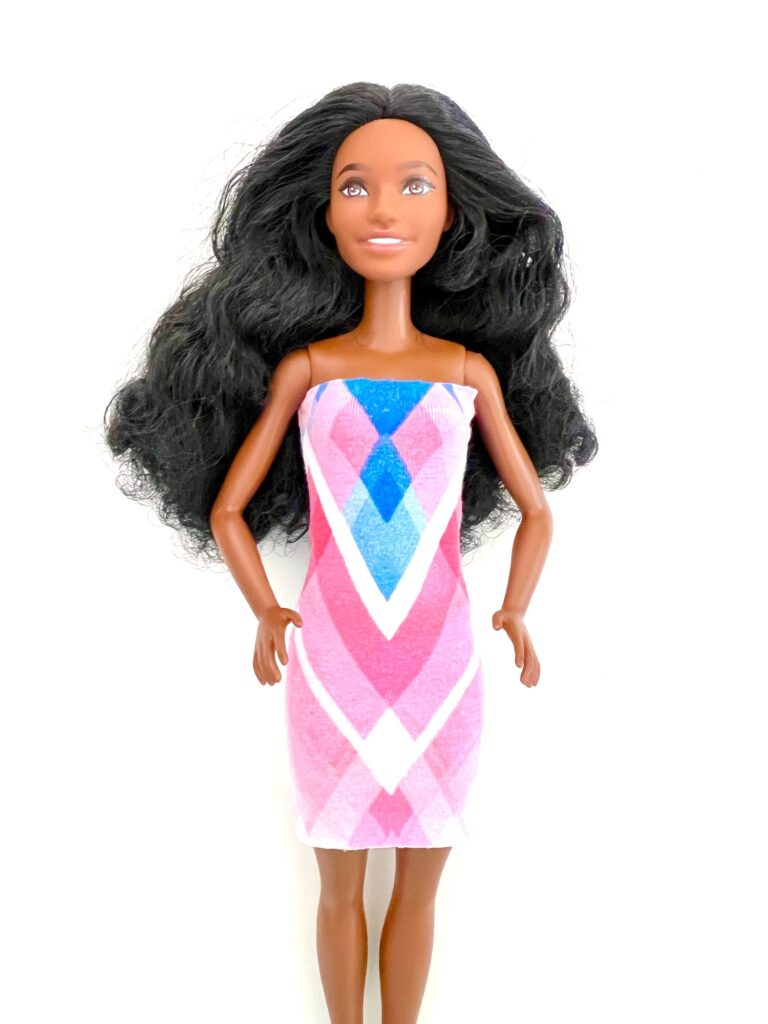 A doll with long black hair, wearing a sleeveless dress featuring pink, white, and blue diamond patterns, stands against a white background.