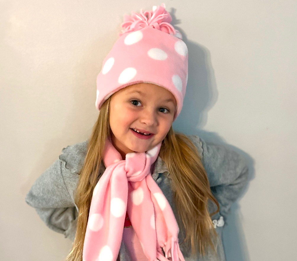 A child with long hair wearing a gray coat, a pink polka dot fleece hat, and matching scarf stands against a plain background, smiling with hands behind their back.
