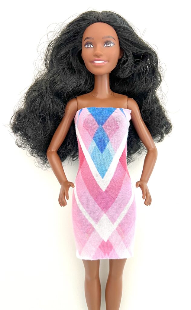 A doll with long, wavy black hair wearing a strapless dress featuring a pink, purple, and blue geometric pattern stands upright against a white background.