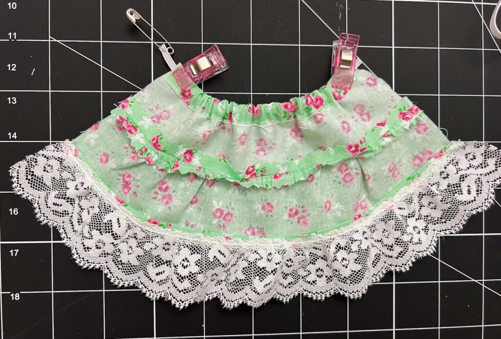 A charming floral fabric skirt with lace trim is clipped onto a grid surface, featuring a softly tiered design.