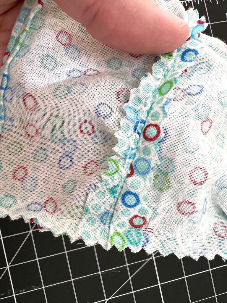 Close-up of a hand holding a piece of white fabric with multicolored circular patterns and zigzag-cut edges, resting on a black grid background.