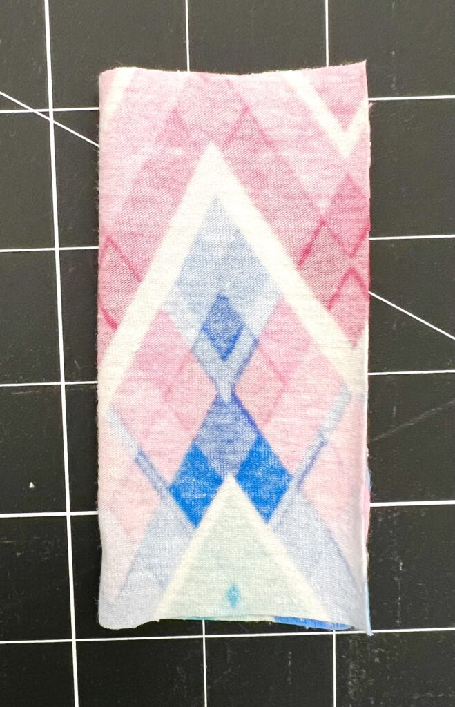 Rectangular fabric swatch with a geometric pattern in shades of pink, blue, and white, placed on a black grid background.