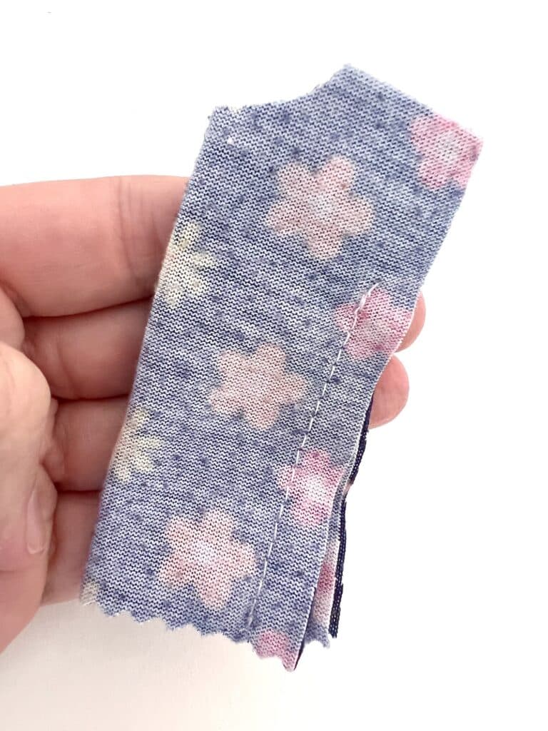 A hand holds a piece of blue fabric with pink and white flowers, zigzag edges, perfect for crafting a whimsical Barbie shirt.