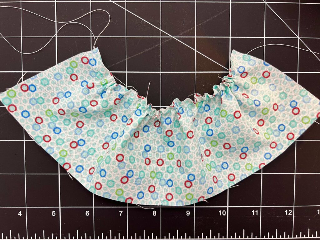 Light blue fabric with multicolored circles is gathered along one edge. Positioned on a black cutting mat with a grid and ruler markings.