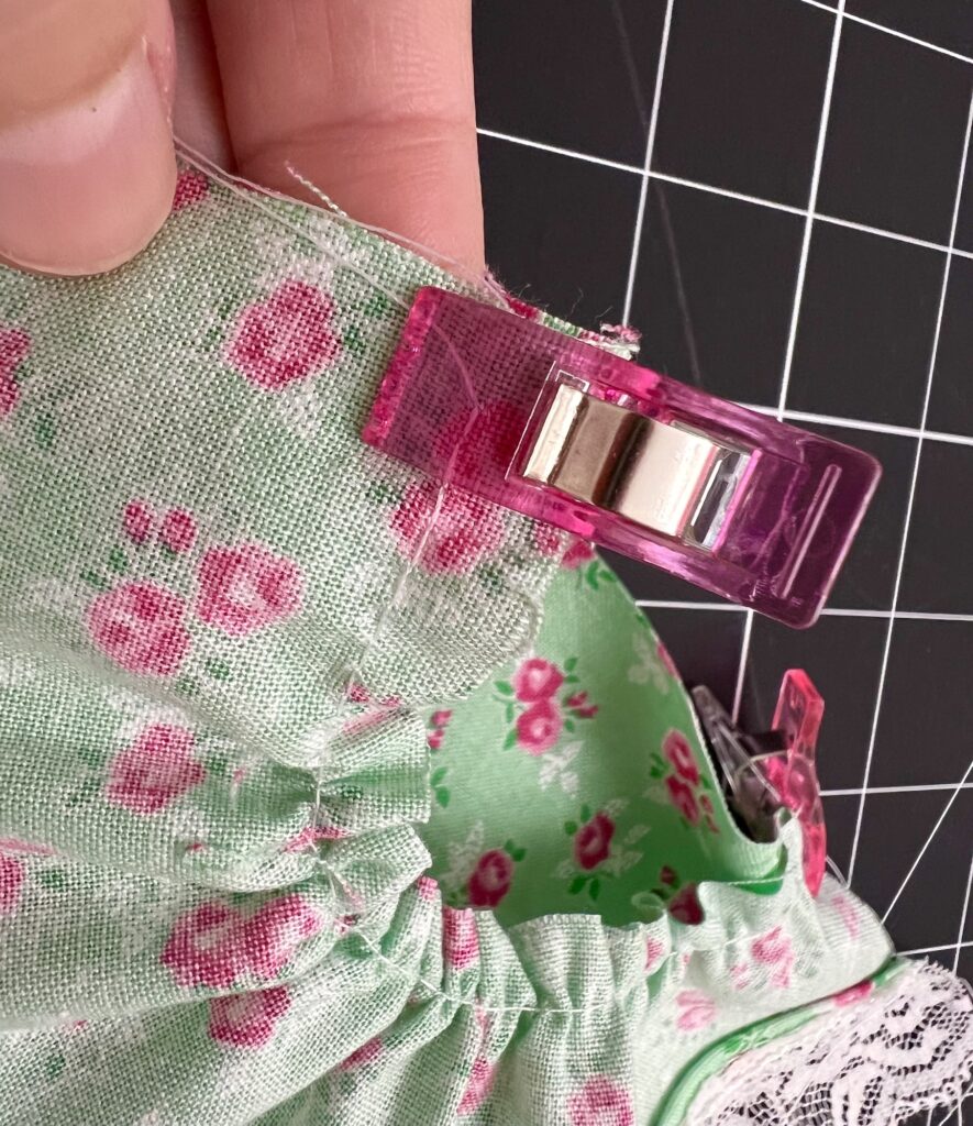 A hand holds a green fabric with pink floral patterns, secured by pink clips, suggesting the beginnings of a tiered skirt. The ruffled edges are partially assembled, set against a black grid background.