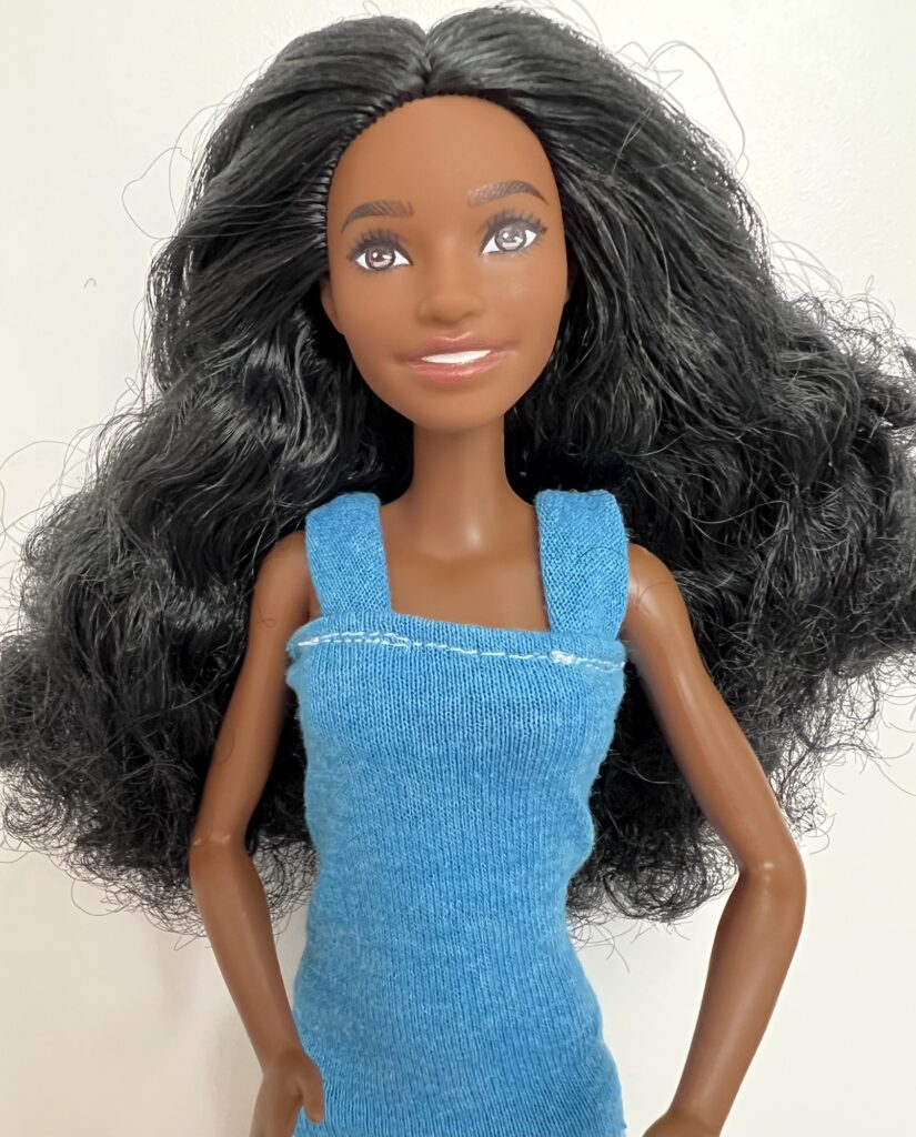 A doll with long black hair wearing a blue dress is standing against a plain white background.