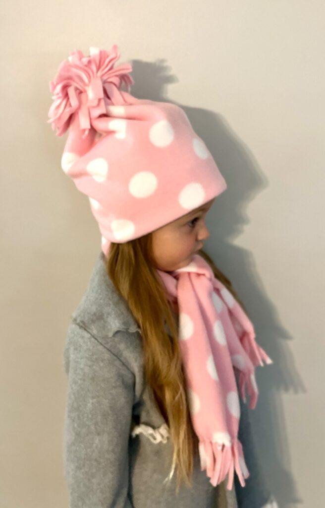 A child wearing a pink fleece hat with polka dots and a pom-pom, along with a matching scarf, stands against a plain wall in a cozy gray coat.