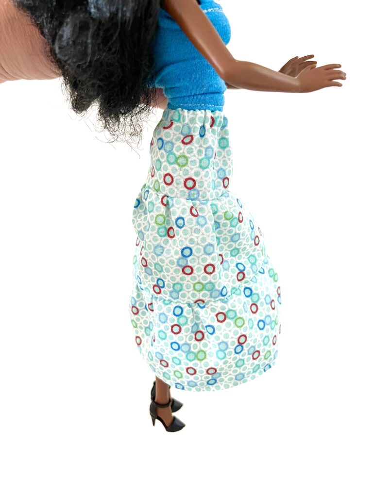 A doll in a blue top and a patterned long skirt with multicolored circles, wearing black high heels.