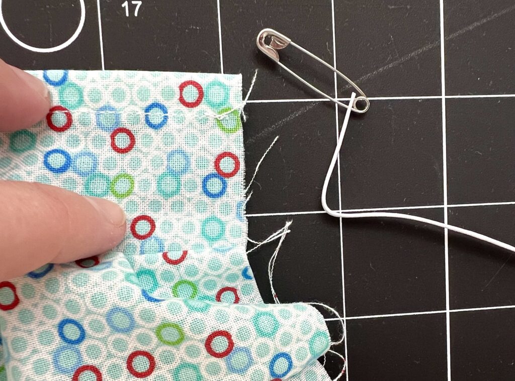 Fingers hold a piece of fabric with circular patterns on a cutting mat. A safety pin is attached to a white string beside the fabric.