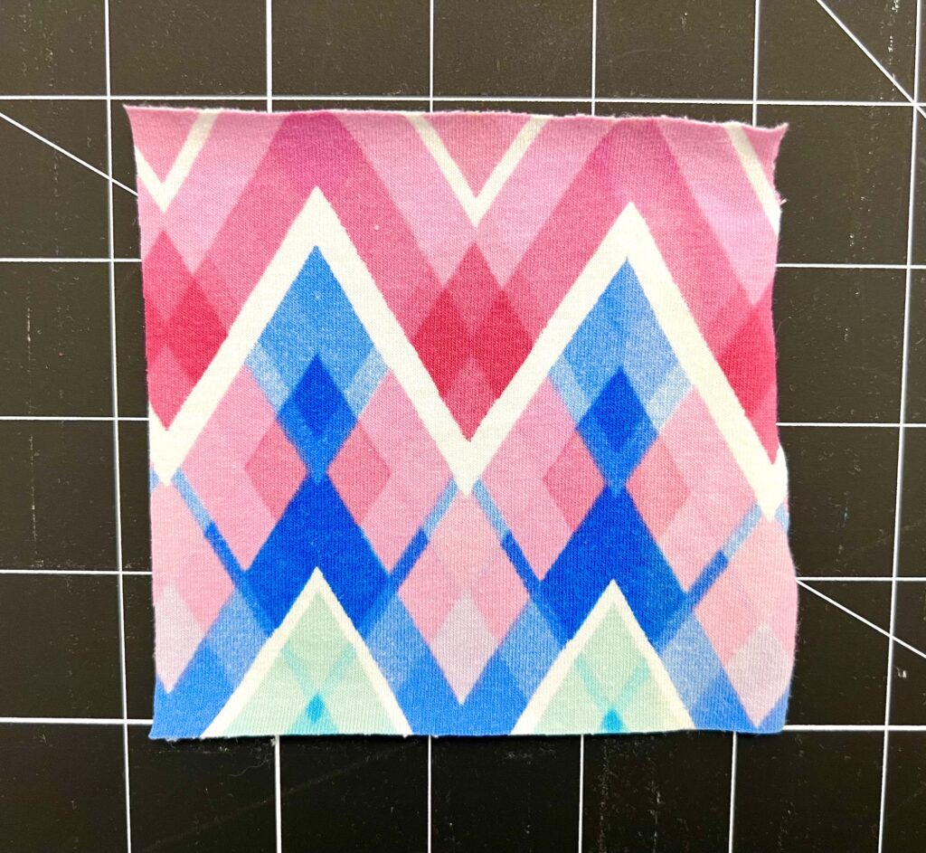 A square piece of fabric with a geometric pattern in pink, blue, and white tones is placed on a black cutting mat with a grid.