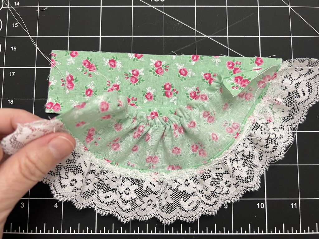 A hand delicately holds fabric adorned with pink floral patterns and white lace trim, envisioning it as a tiered skirt masterpiece on a black grid cutting mat.