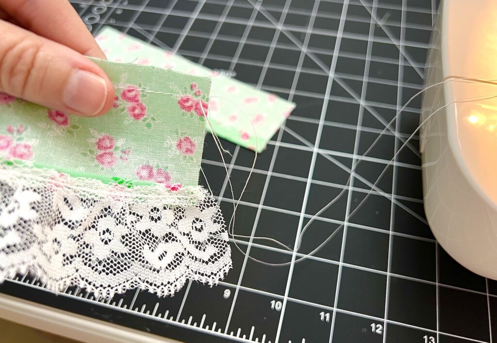 A hand holds a piece of green floral fabric with white lace trim, perfect for creating a tiered skirt, above a cutting mat next to a sewing machine.