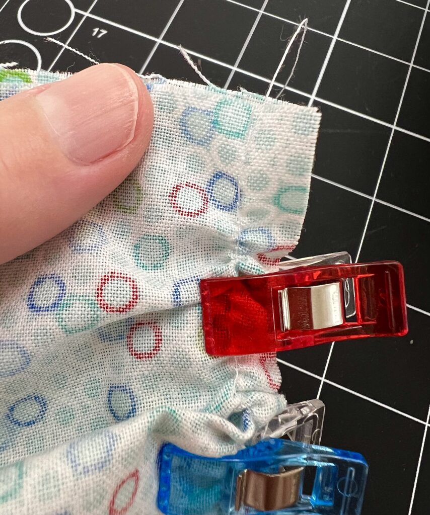 Fingers holding fabric with multicolored dots, secured by red and blue clips, over a black grid cutting mat.