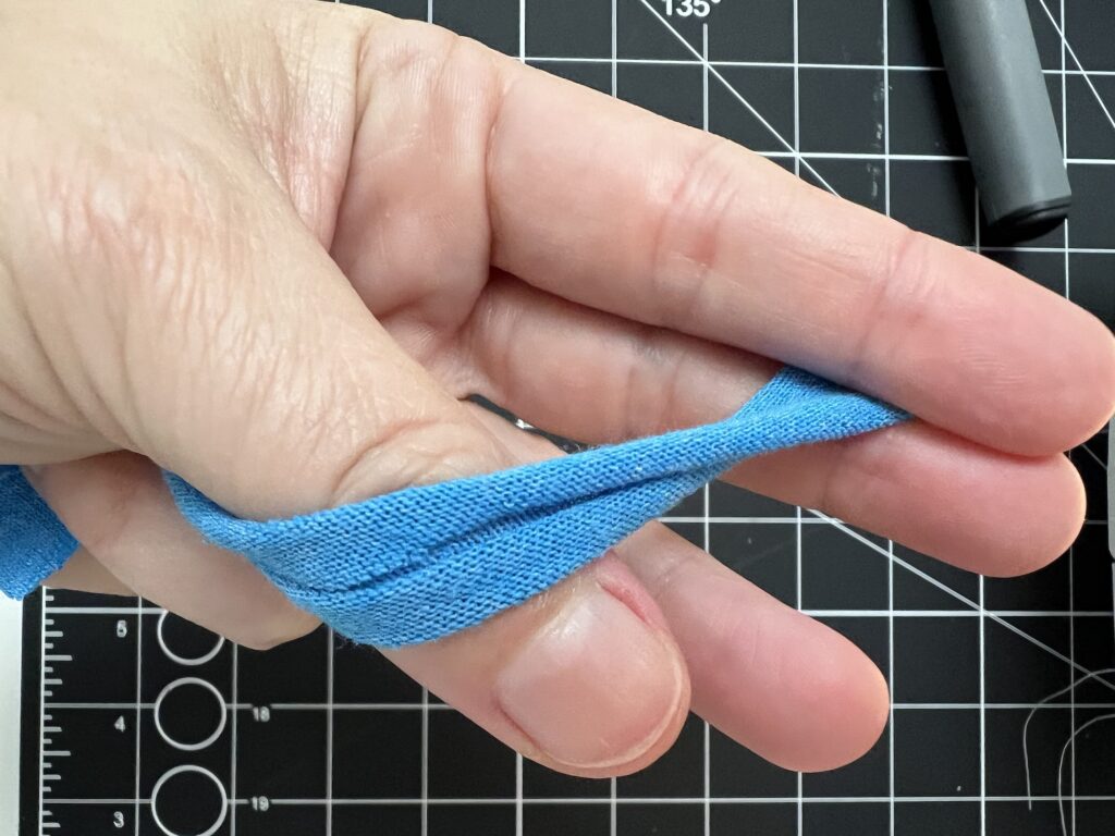 A hand holding a blue fabric strip over fingers, with a grid cutting mat in the background.