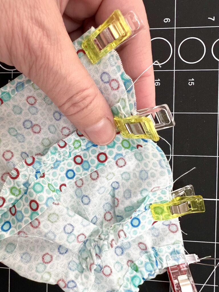 Close-up of a hand holding patterned fabric pieces clipped together with multi-colored clips on a black cutting mat.