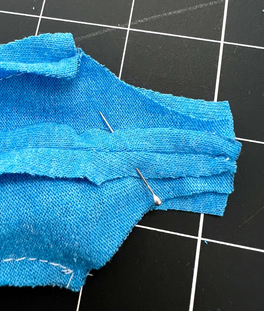 A close-up of blue fabric pieces pinned together on a grid-patterned cutting mat, showcasing the intricate sewing details of a swimsuit in progress.