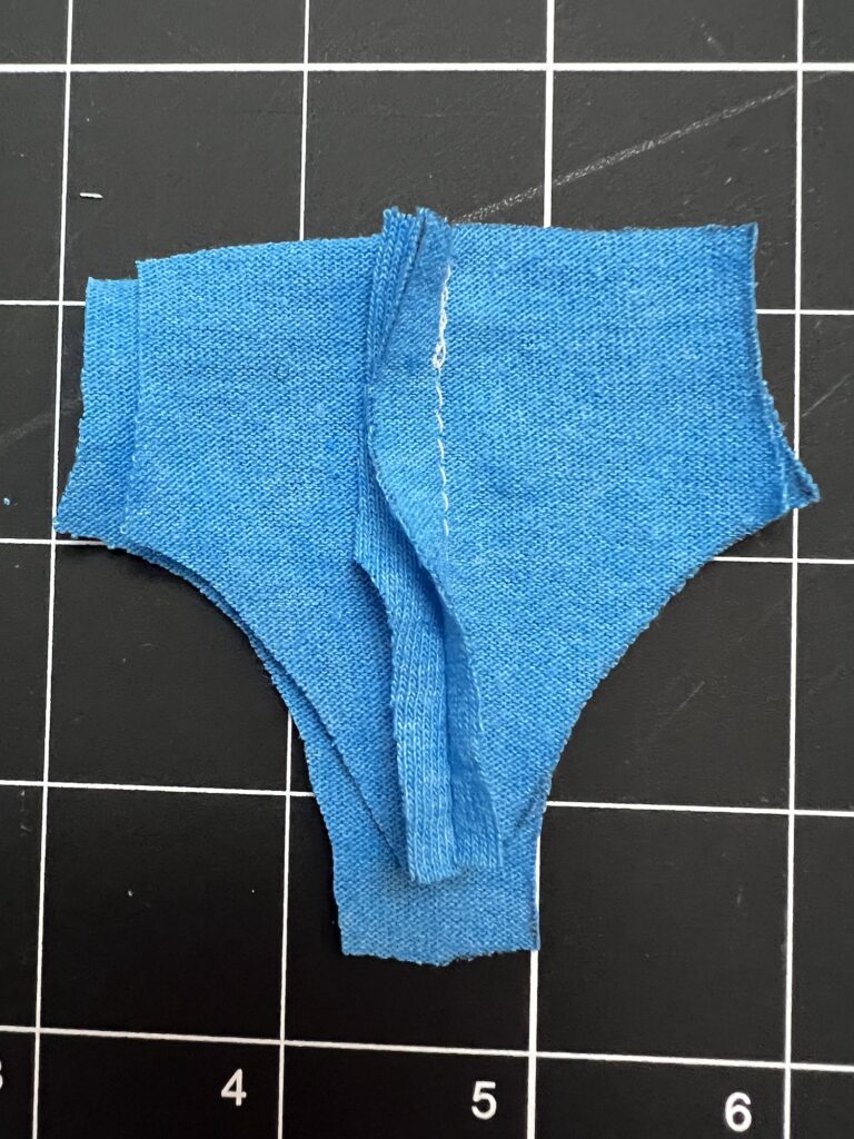 A small, blue, unfinished fabric piece resembling shorts is placed on a black grid cutting mat.