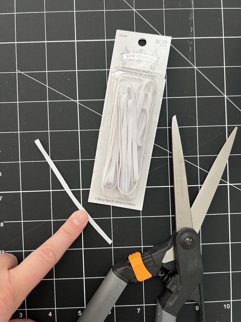 Package of white elastic, a piece cut, scissors, and a finger pointing at it, on a black grid cutting mat.