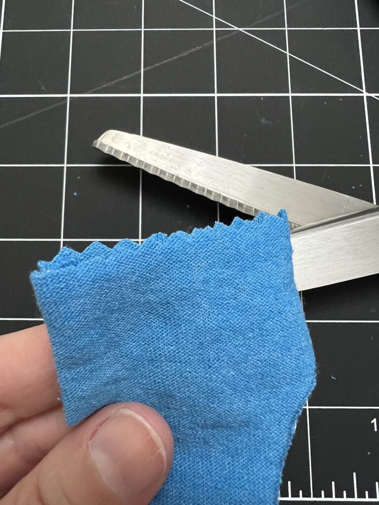 A hand holds a piece of blue fabric with zigzag edges. Scissors with a zigzag blade are in the background on a grid cutting mat.