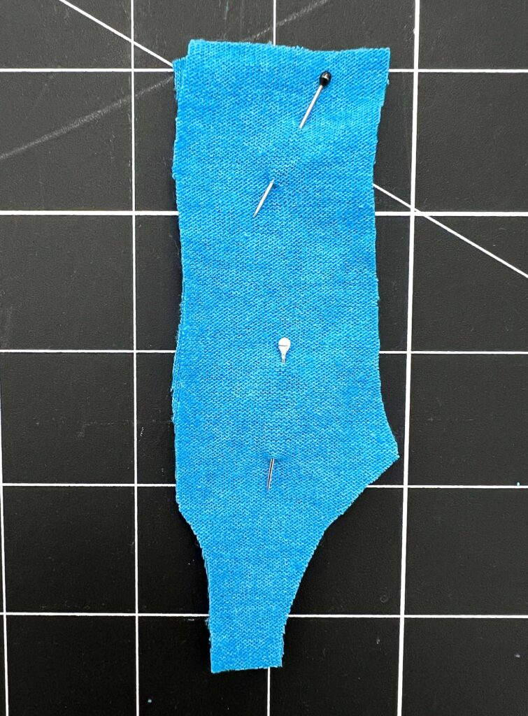 A piece of blue fabric, reminiscent of a new swimsuit in the making, is pinned onto a black and white grid cutting mat.