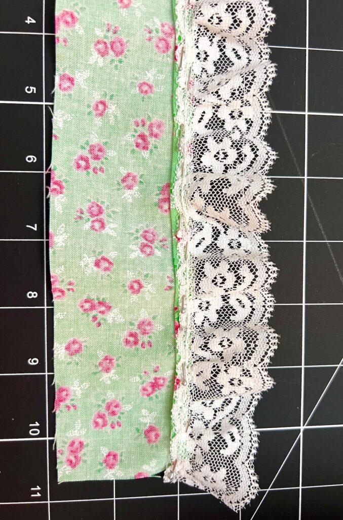 Two fabric strips rest on a black grid mat: one is a green piece adorned with pink floral patterns, while the other is delicate white lace, ideal for crafting a tiered skirt.