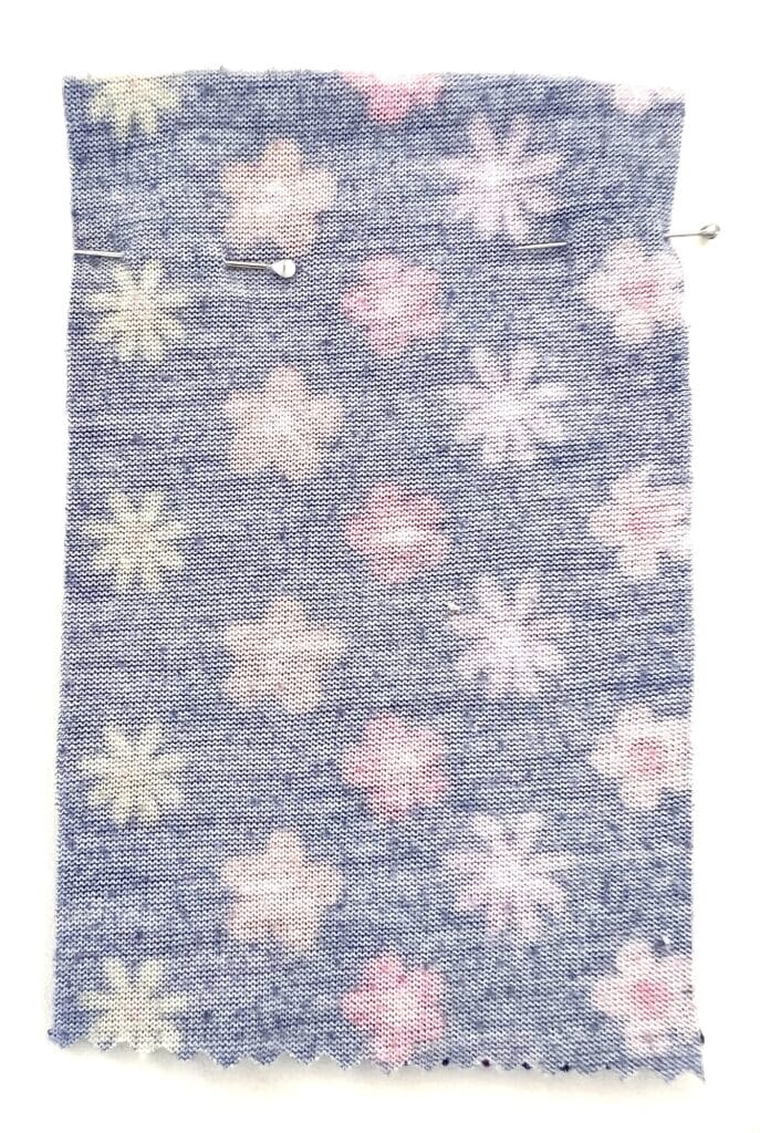 A rectangular piece of blue textured fabric with pink and white floral patterns, reminiscent of a Barbie shirt, secured with two pins.