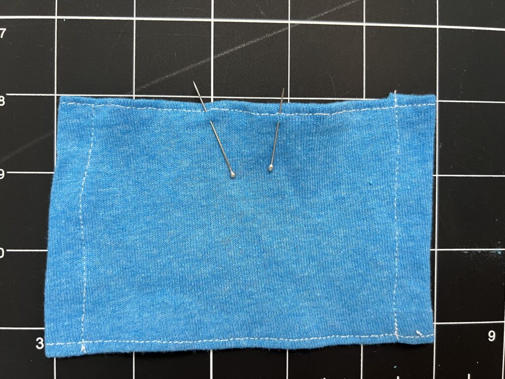 A rectangular piece of blue fabric with white stitching is placed on a black grid surface. Two needles are inserted near the center.