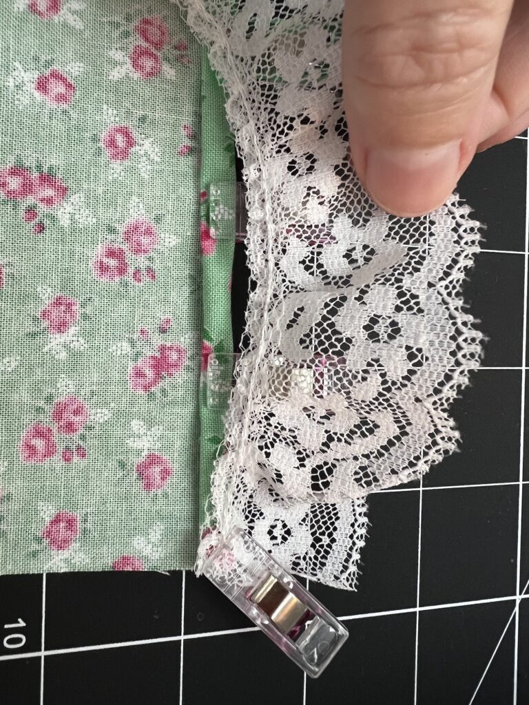 A hand holds lace fabric clipped to floral fabric on a grid cutting mat, ready to be transformed into an elegant tiered skirt.