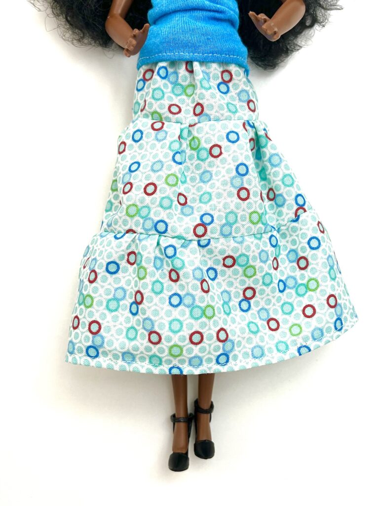 A doll wearing a blue sleeveless top and a long skirt with a light blue, white, and red polka dot pattern. The doll has black shoes.