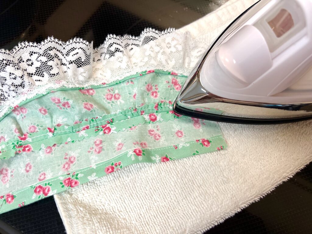 An iron is pressing a green floral fabric on a white towel with lace edging, reminiscent of the delicate charm found in a tiered skirt.