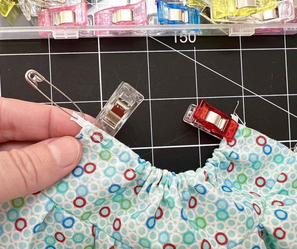 A hand holds fabric with a colorful pattern, secured by clips and a safety pin, on a black grid cutting mat. More clips are visible in a container above.