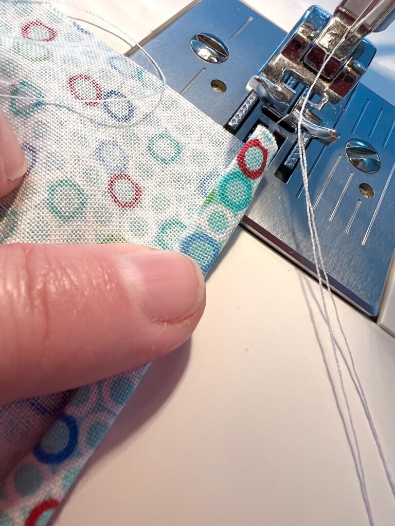 Close-up of a person's hand guiding fabric with a colorful circular pattern under a sewing machine's presser foot, with white thread visible.