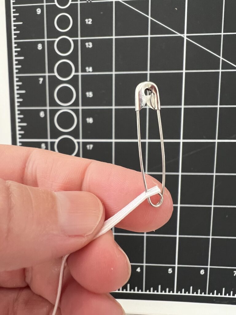 A hand holding a safety pin attached to a white elastic band in front of a grid cutting mat.