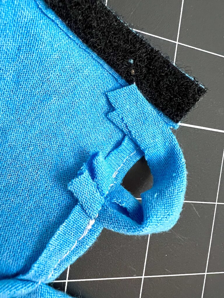 Close-up of a blue fabric piece with a cutout loop and Velcro strap on a black grid background.