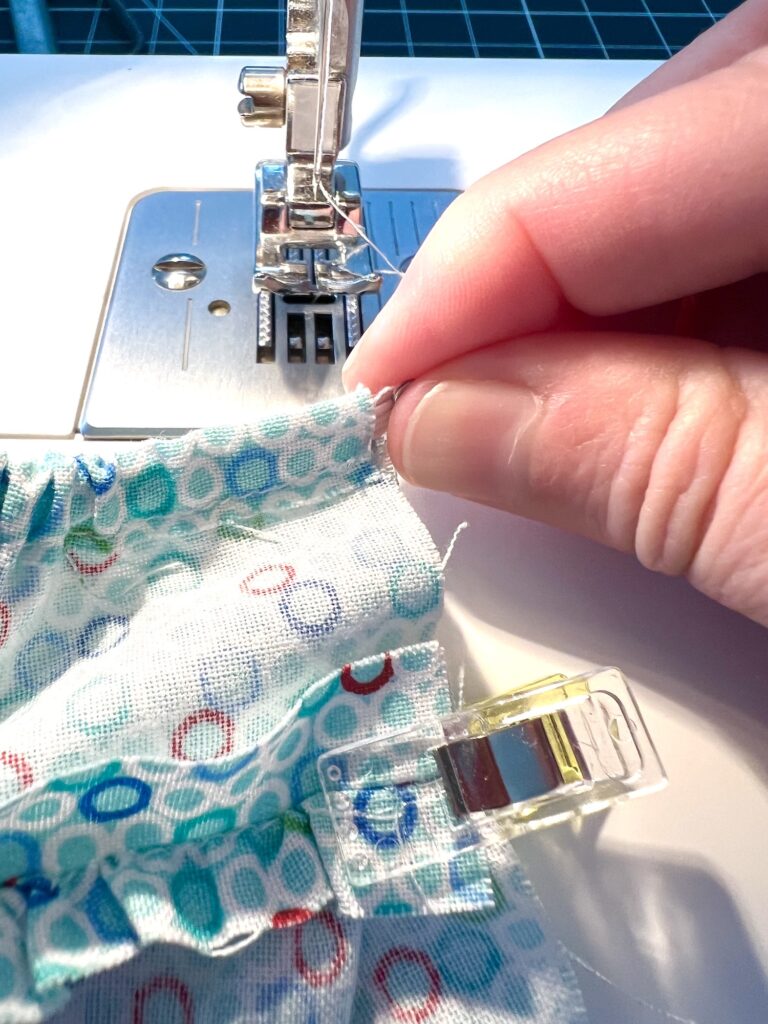 Close-up of a person sewing pleated fabric with a sewing machine. A hand adjusts the fabric, which is secured with a plastic clip.