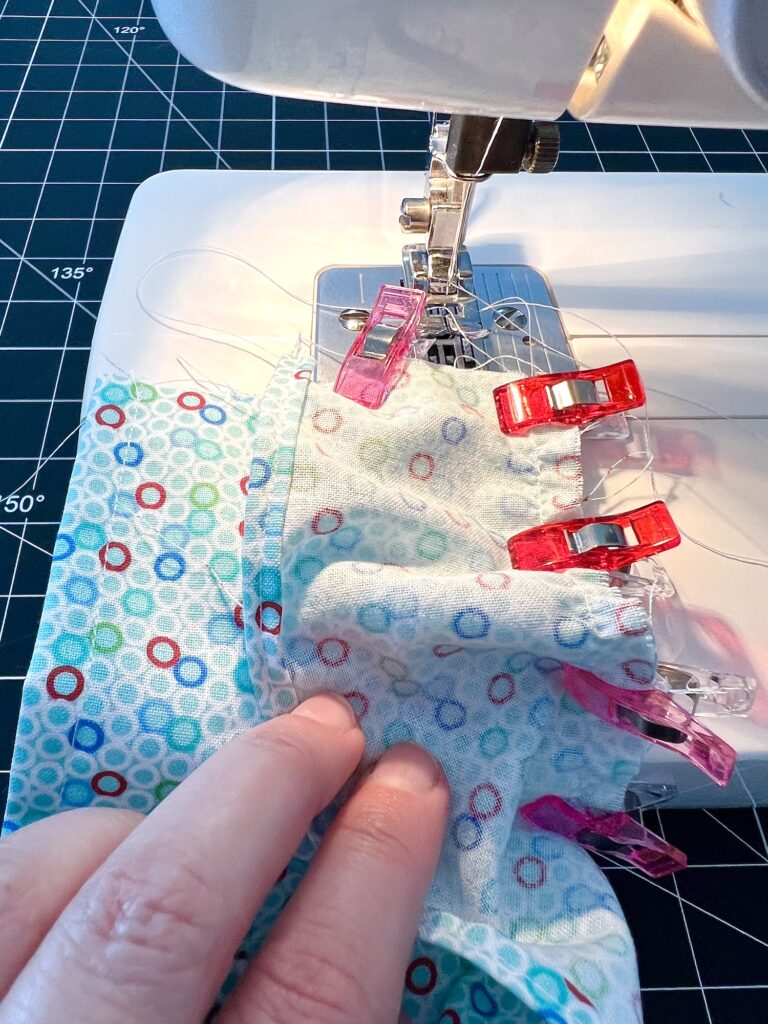 A person sewing fabric with a pattern of multicolored circles on a sewing machine, held together by red clips, on a grid cutting mat.