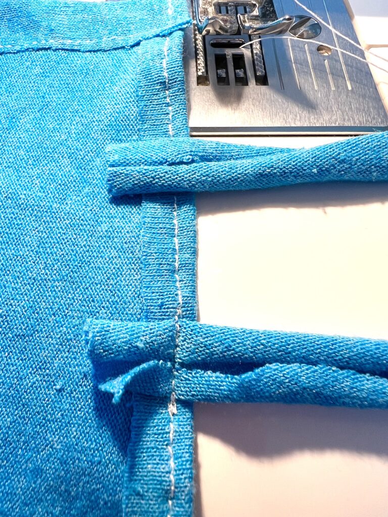 A sewing machine stitches a blue fabric with white thread, showing an unfinished edge and seam.