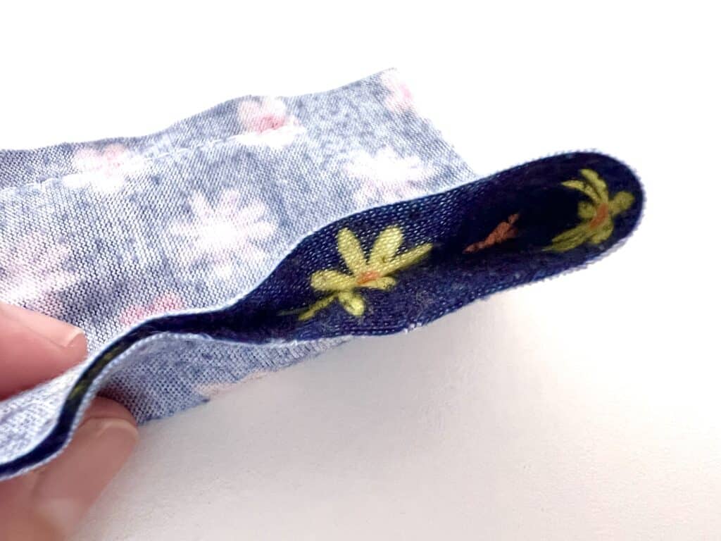 Close-up of a finger holding blue fabric with white and pink flowers, reminiscent of a classic Barbie shirt. The reverse side reveals navy fabric adorned with yellow flowers and green leaves.