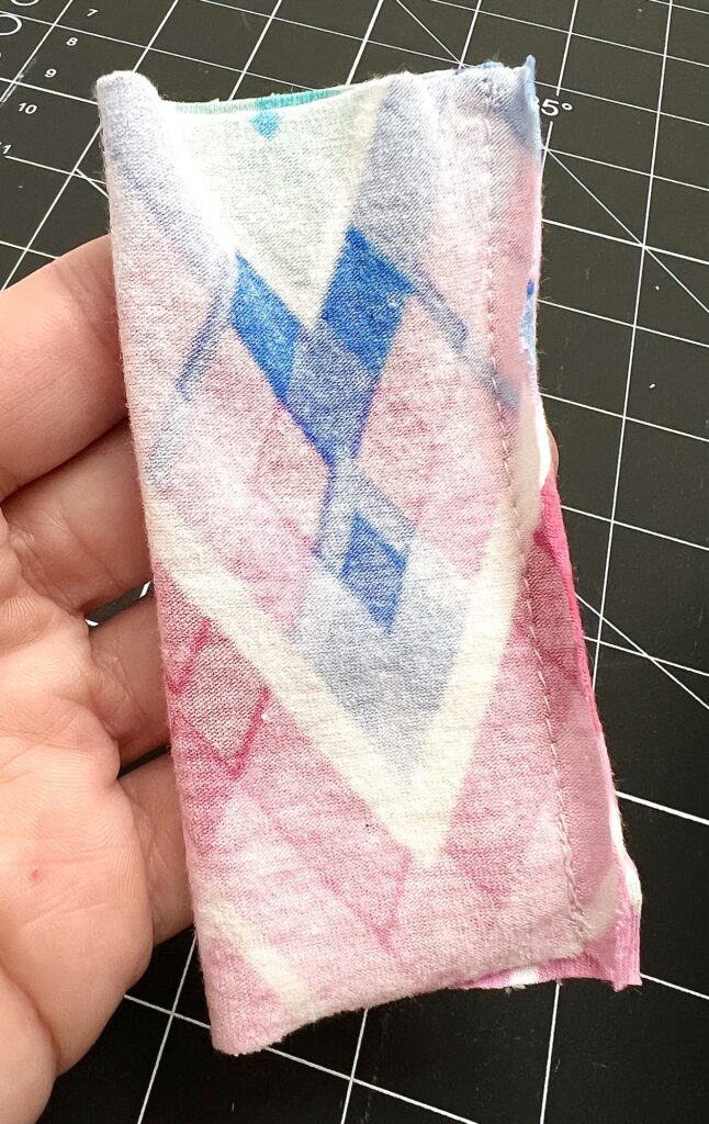 A hand holding a folded piece of fabric with a geometric pattern in pink, blue, and white tones. The background features a cutting mat with a grid design.