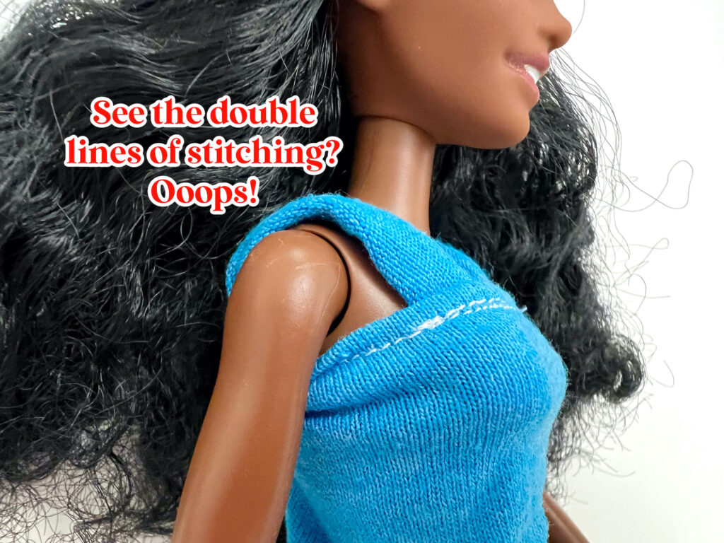 A doll with long dark hair wears a blue top with noticeable double lines of stitching. Text reads, "See the double lines of stitching? Ooops!.